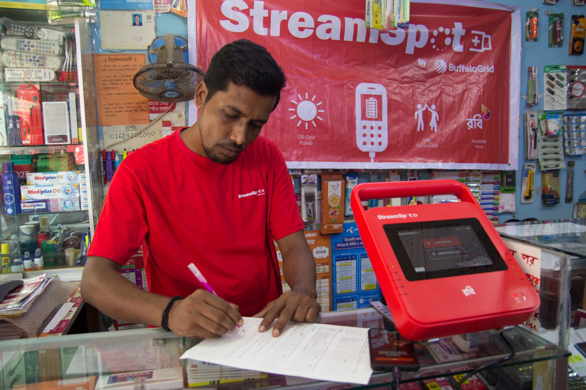 Shopkeeper writing at a counter beside a red BuffaloGrid hub and StreamSpot banner