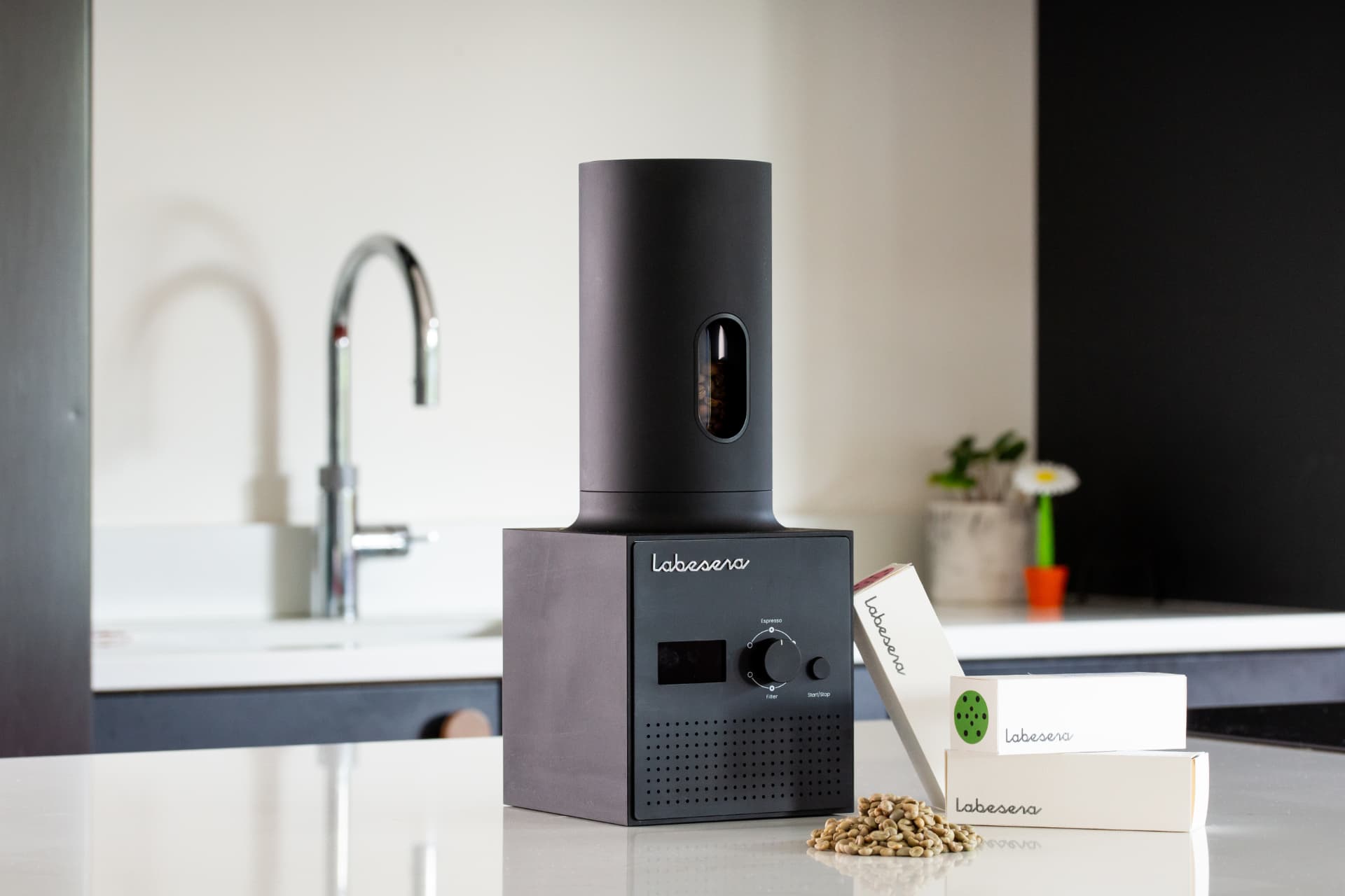 Labesera coffee roaster in matt black on a white kitchen counter, with green beans and Labesera coffee pouches beside it